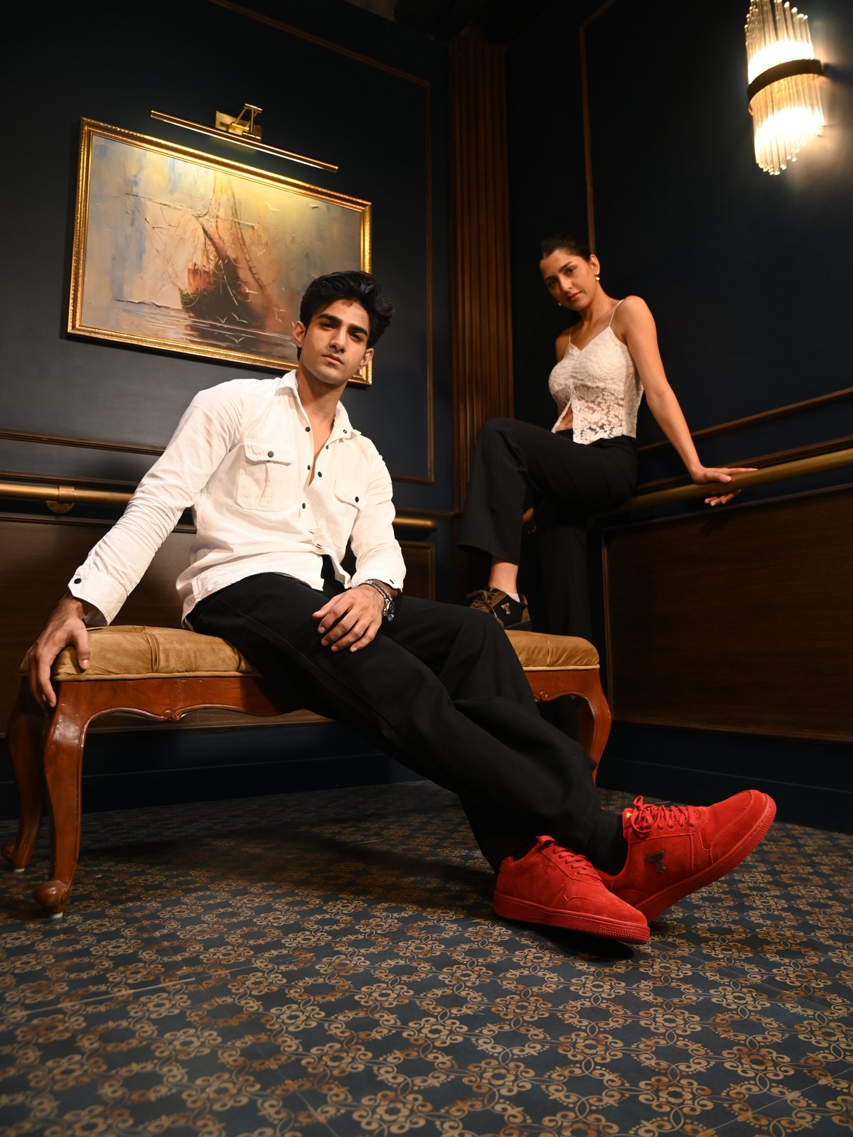 Man in a white shirt reclining on a bench in red suede sneakers, a woman behind him