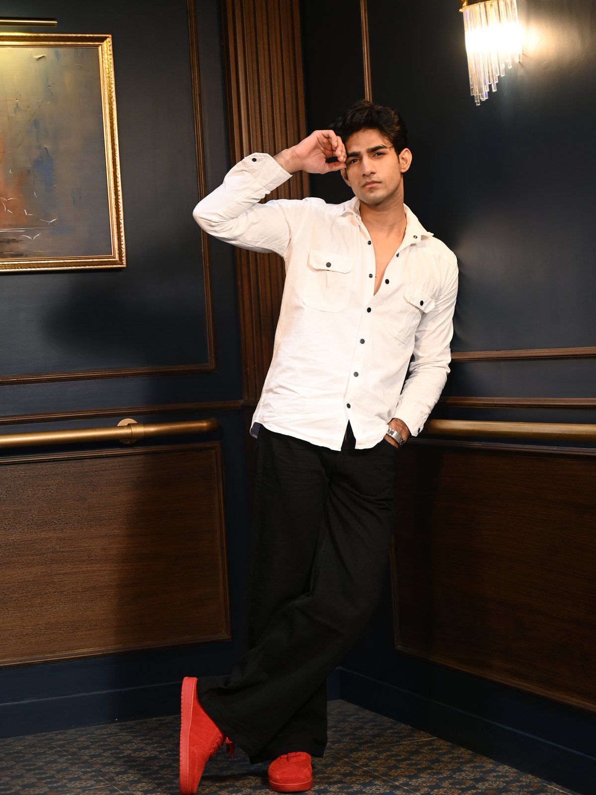 Man in a white shirt leaning on a wall rail in red suede sneakers, hand to his head