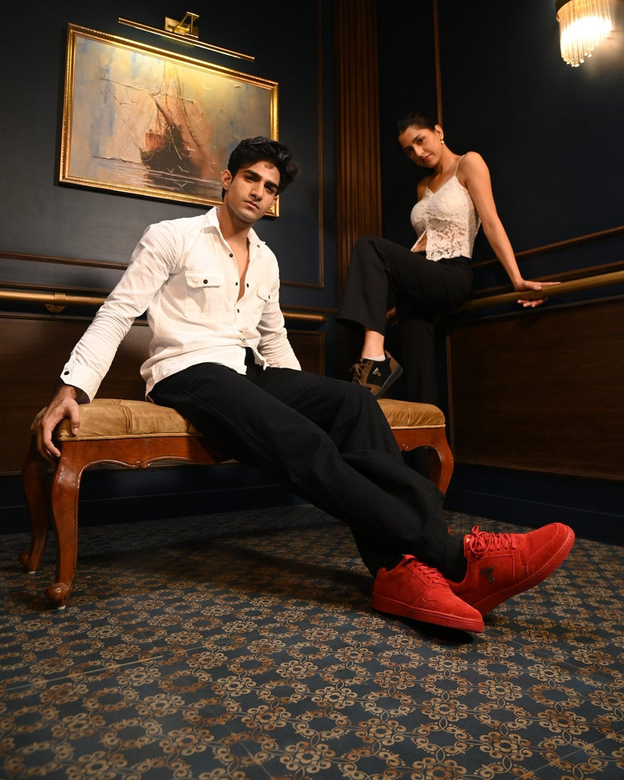 Man in a white shirt reclining in red suede THYLACIN sneakers, a woman seated behind him