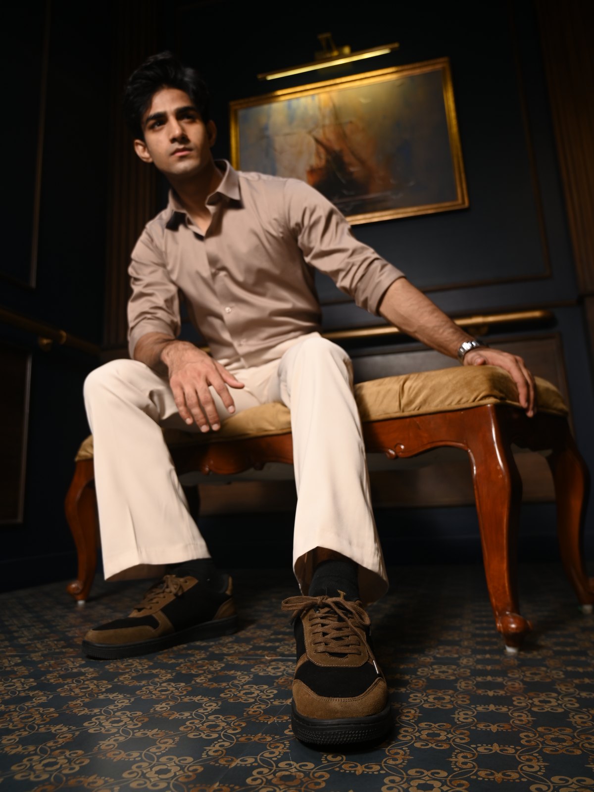 Man in cream trousers seated on a velvet bench, wearing The Copper Jackal tan-and-black sneakers