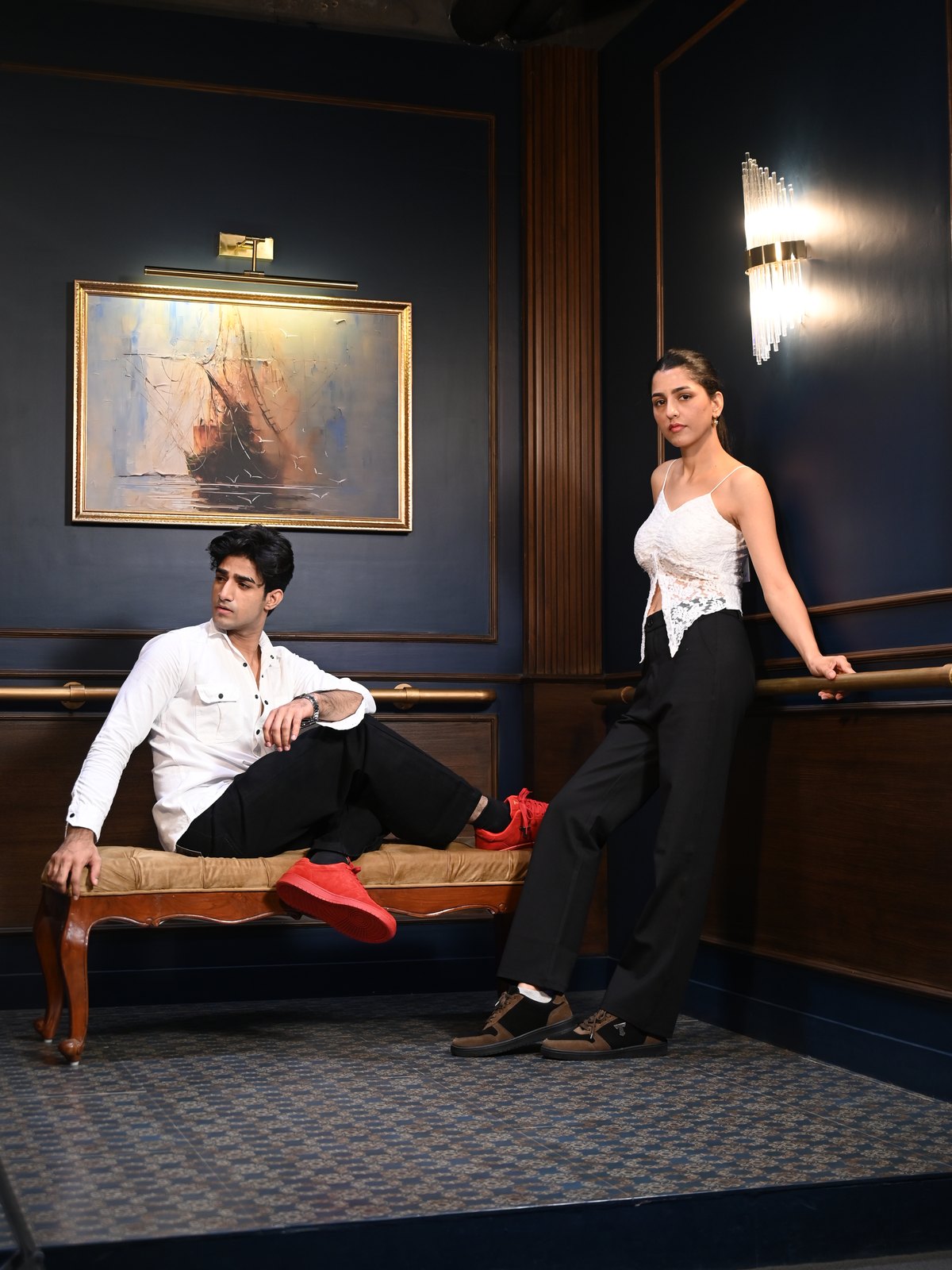 Couple in a dark study: he reclines on a bench in red suede sneakers, she leans on the rail
