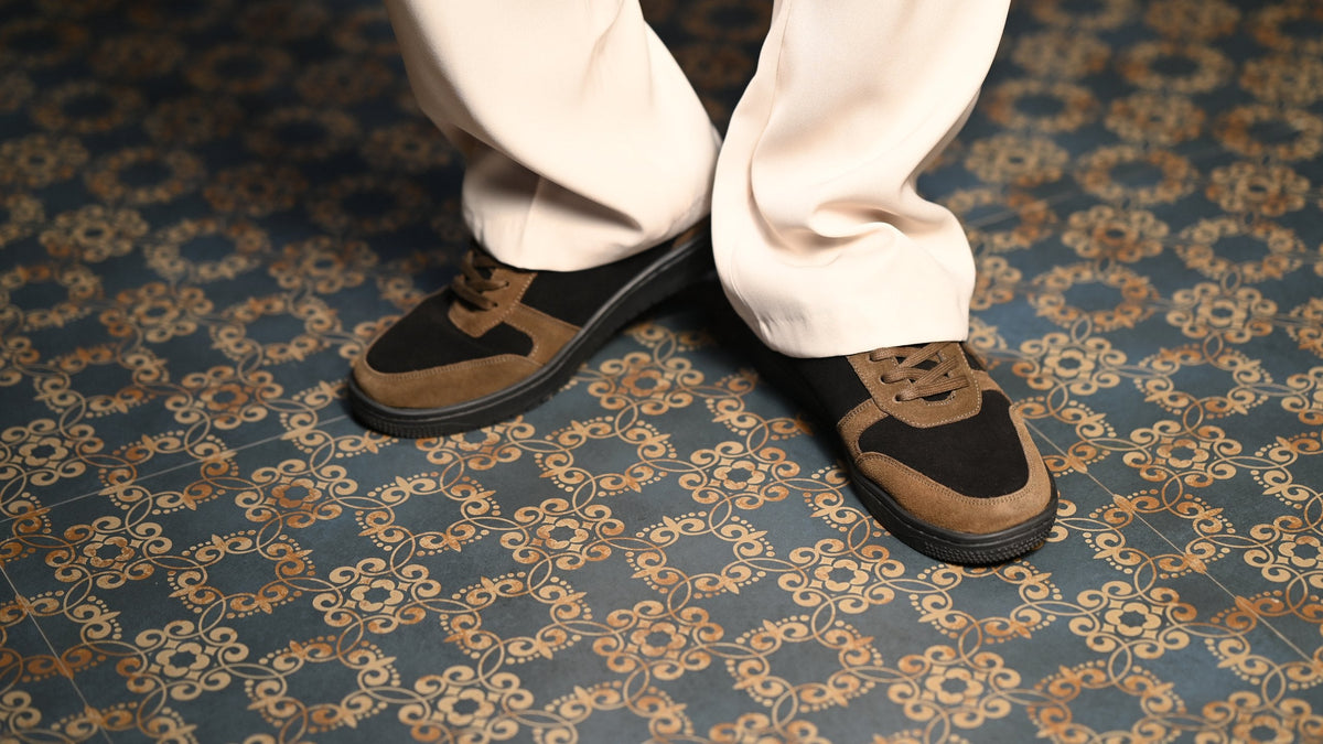 The Copper Jackal tan-and-black sneakers with cream trousers on a patterned carpet
